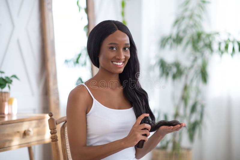 Cosmetics and healthy beautiful long hair. Smiling young african american female takes care of hair. Applying thermal protect hair spray, with smooth hair