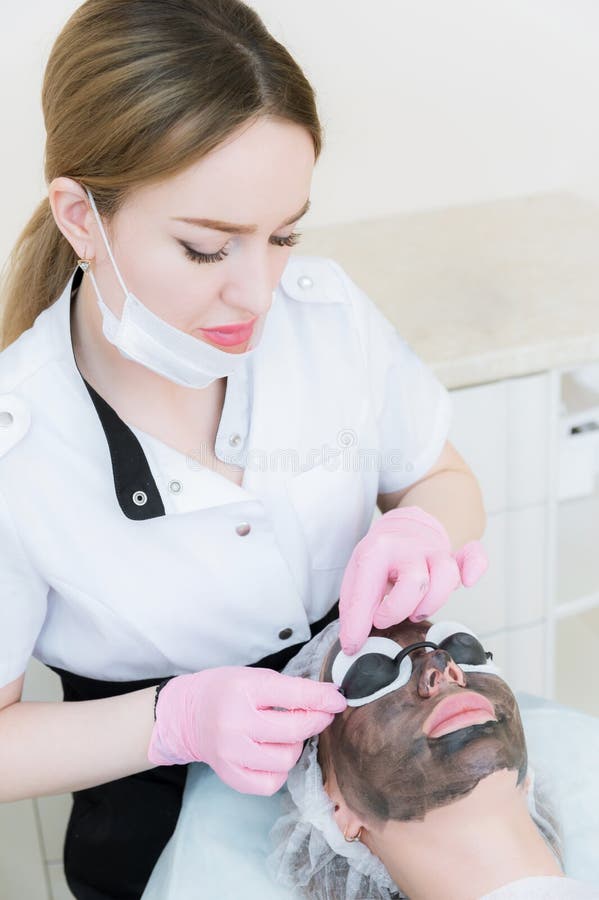 The cosmetologist in pink gloves with a brush applies a carbon mask for peeling on the face of a young girl in a stock image The cosmetologist in pink gloves with a brush applies a carbon mask for peeling on the face of a young girl in a stock image