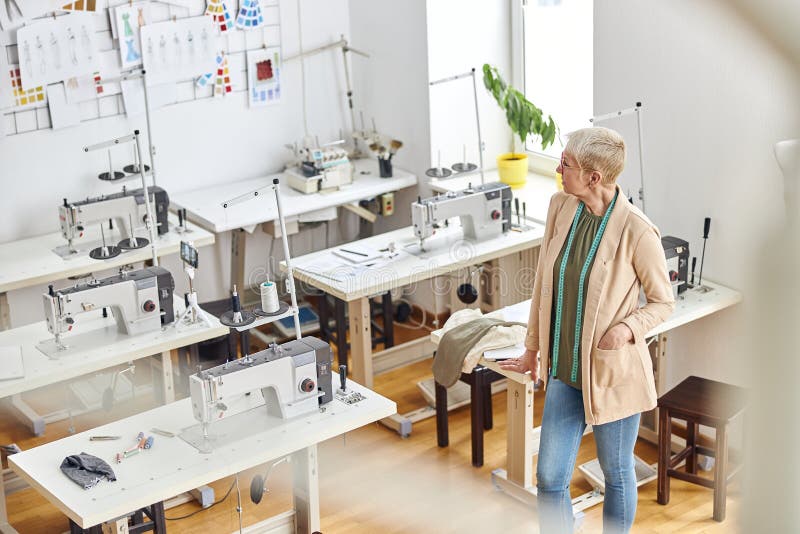 Mature lady dressmaker with stylish short haircut and tape measure stands in empty sewing workshop equipped with contemporary machines. Mature lady dressmaker with stylish short haircut and tape measure stands in empty sewing workshop equipped with contemporary machines