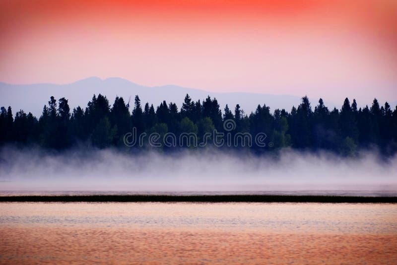 Coucher du soleil de lever de soleil sur le lac avec la brume se levant des pins et des montagnes de l'eau à l'arrière-plan photo stock Coucher du soleil de lever de soleil sur le lac avec la brume se levant des pins et des montagnes de l'eau à l'arrière-plan photo stock