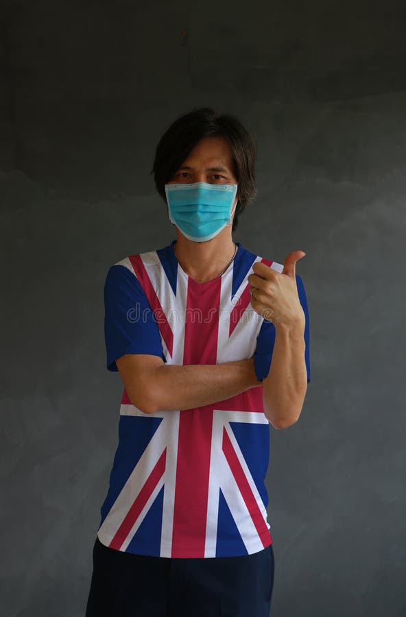 Masked man wearing Union Jack flag color of shirt and cross one s arm with thumbs up on dark wall background. Concept of protection and fighting COVID. Masked man wearing Union Jack flag color of shirt and cross one s arm with thumbs up on dark wall background. Concept of protection and fighting COVID