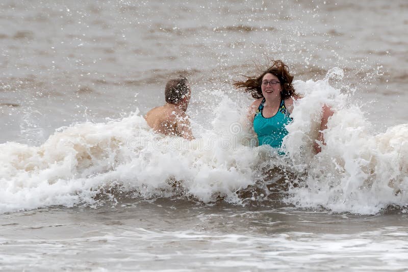 Saint John, NB, Canada - January 1, 2020: A man and woman participate in the annual `polar bear swim` at Mispec Beach. The woman faces the camera while a wave crashes into her back. Saint John, NB, Canada - January 1, 2020: A man and woman participate in the annual `polar bear swim` at Mispec Beach. The woman faces the camera while a wave crashes into her back