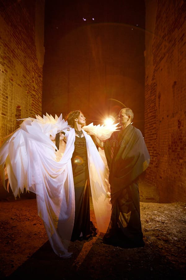 Couple in dark clothes in an abandoned castle with old brick walls. Man in black dress and woman with white wings posing