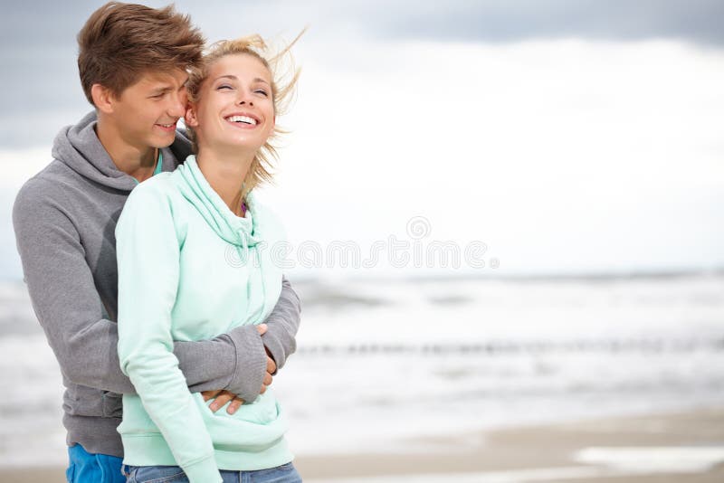 Couple embracing and having fun wearing warm clothes outside on coast behind blue sky