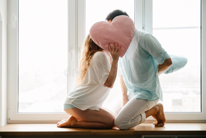 Couple girl and guy play with pillows near window.