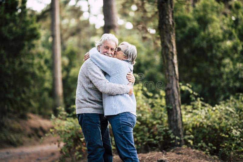 Couple of happy and cheerful people caucasian senior hug with love and smile with defocused green forest in background - outdoor