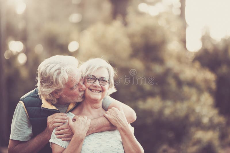 Couple of happy and cheerful people caucasian senior hug with love and smile with defocused green forest in background - outdoor