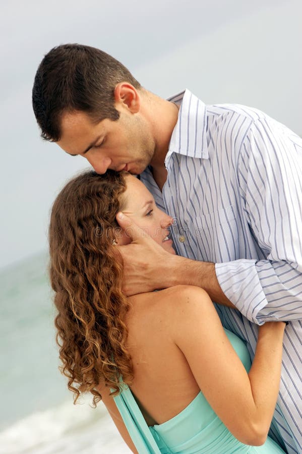 A young woman is being held close by a man, He is kissing the top of her head. With ocean waves in the background. A young woman is being held close by a man, He is kissing the top of her head. With ocean waves in the background.