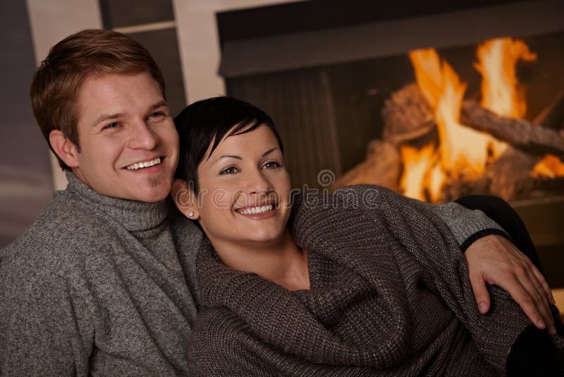 Young couple hugging in front of fireplace at home, looking away, smiling. Young couple hugging in front of fireplace at home, looking away, smiling.