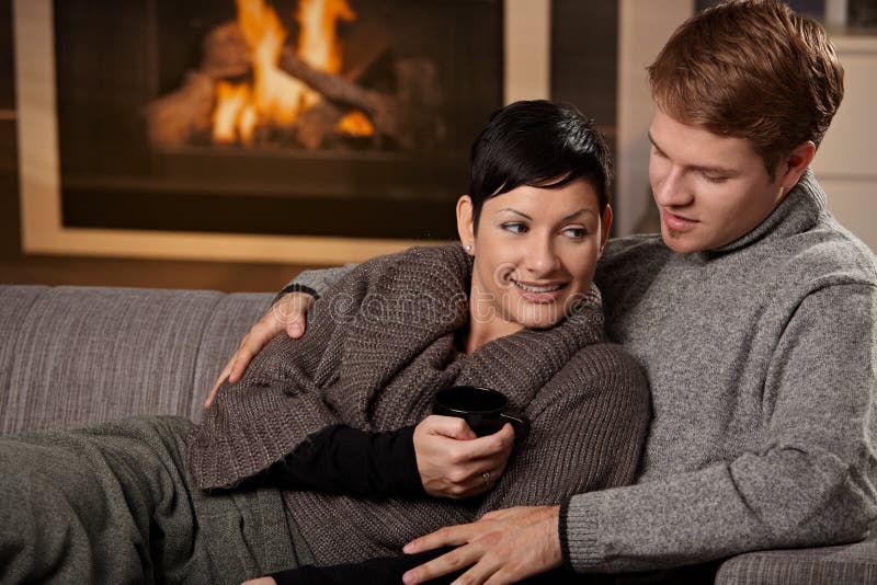 Young couple hugging on sofa in front of fireplace at home, smiling. Young couple hugging on sofa in front of fireplace at home, smiling.