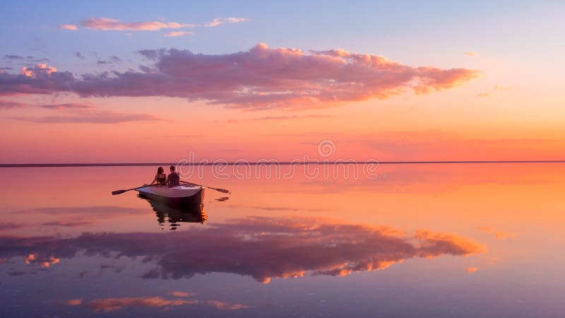 A couple in love look at beautiful sunset in a rowing boat on the lake. Pink sky and vanilla clouds. Romantic scene - lovers ride a boat in nature during sunset. Amazing landscape with people.