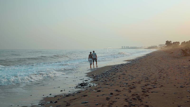 A couple, a man and a woman, walk hand in hand along a sandy beach barefoot on the incoming waves at sunset on a summer