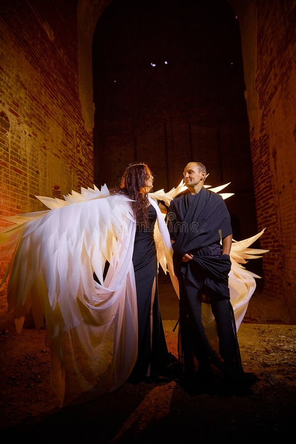 Couple in old dark clothes in abandoned castle with old brick walls. Man in black dress and woman with white wings