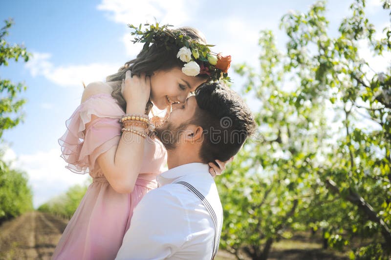 Close-up couple portrait of a girl and guy looking for a wedding dress, a pink dress flying with a wreath of flowers on her head on a background garden and the blue sky, and they hug and pose