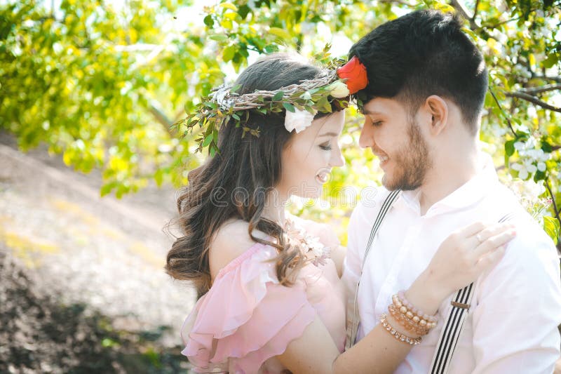 Close-up couple portrait of a girl and guy looking for a wedding dress, a pink dress flying with a wreath of flowers on her head on a background garden and the blue sky, and they hug and pose