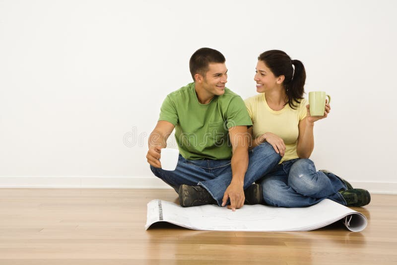 Attractive couple sitting on home floor with coffee cups looking at house plans and smiling at eachother. Attractive couple sitting on home floor with coffee cups looking at house plans and smiling at eachother.
