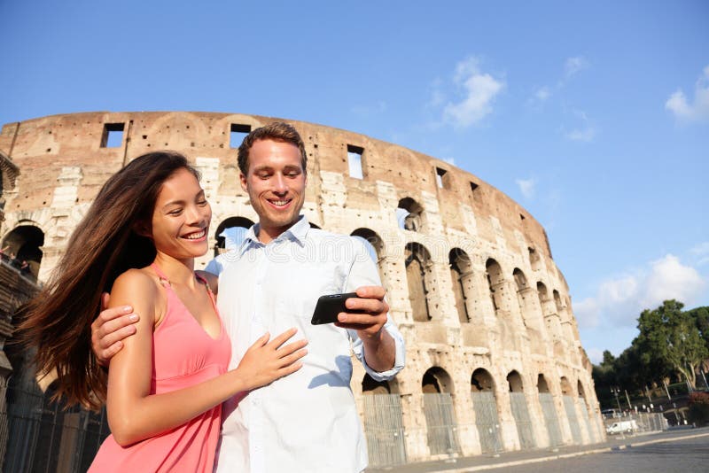 Couple in Rome by Colosseum using smart phone looking at pictures or using travel app in Italy. Happy lovers on honeymoon sightseeing Coliseum. Love and travel concept with multiracial couple.