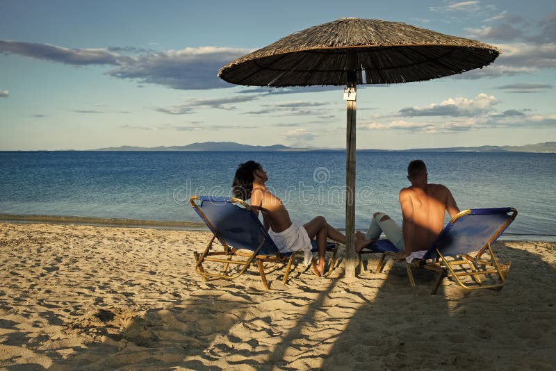 Couple secrets fantasy. Couple in love at sea resort sits on loungers under umbrella on sand beach. Couple on vacation royalty free stock images Couple secrets fantasy. Couple in love at sea resort sits on loungers under umbrella on sand beach. Couple on vacation royalty free stock images