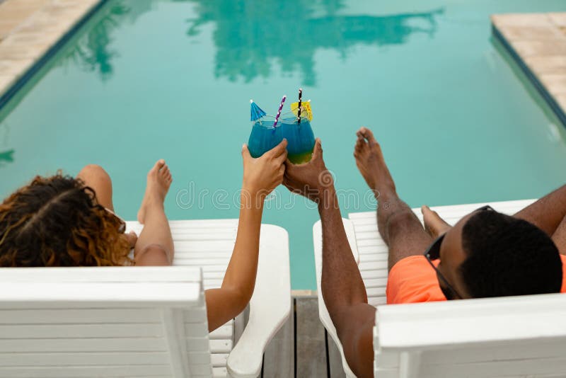 Couple toasting glasses of cocktail while relaxing on a sun lounger near swimming pool at the backya