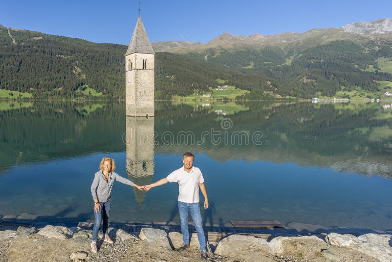 couple tourists pose front old bell tower curon venosta which perfectly reflected still water lake 197851401