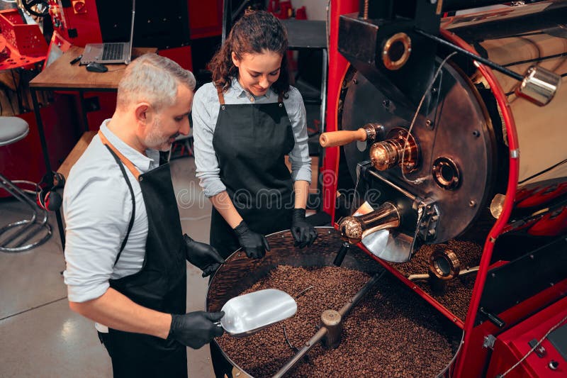 Couple of two baristas checking the quality of the coffee beans standing with scoop near the roaster machine at the roastery. High