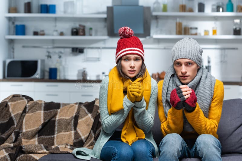 Couple in warm hats and gloves. Freezing couple in warm hats and gloves looking at camera while sitting on sofa in cold kitchen