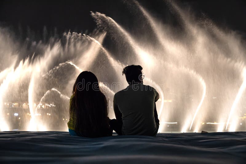 Dubai, UAE - May 25, 2018: A couple watching the light and water show of Dubai Mall fountains from a floating platform after dark. Dubai, UAE - May 25, 2018: A couple watching the light and water show of Dubai Mall fountains from a floating platform after dark.