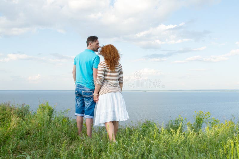 Couples attrayants étreignant et regardant le lac et le ciel bleu avec des nuages le coucher du soleil Couples dans l'herbe images libres de droits Couples attrayants étreignant et regardant le lac et le ciel bleu avec des nuages le coucher du soleil Couples dans l'herbe images libres de droits