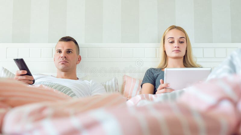 Couples caucasiens attrayants dans le lit Équipez TV de observation, Web de lecture rapide de femme sur le comprimé numérique Cou image stock Couples caucasiens attrayants dans le lit Équipez TV de observation, Web de lecture rapide de femme sur le comprimé numérique Cou image stock
