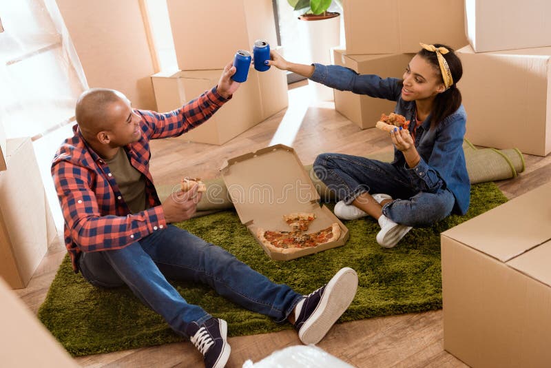 couples d'afro-américain mangeant de la pizza et tintant avec des boîtes de soude en nouvel appartement photo libre de droits couples d'afro-américain mangeant de la pizza et tintant avec des boîtes de soude en nouvel appartement photo libre de droits