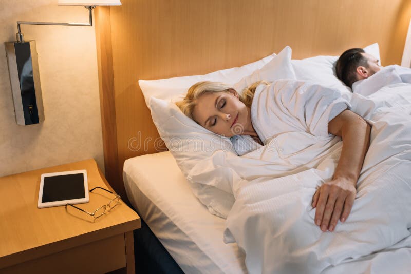 Couples mûrs dans des peignoirs dormant dans le lit et le comprimé numérique avec des lunettes sur la table dans l'hôtel photographie stock