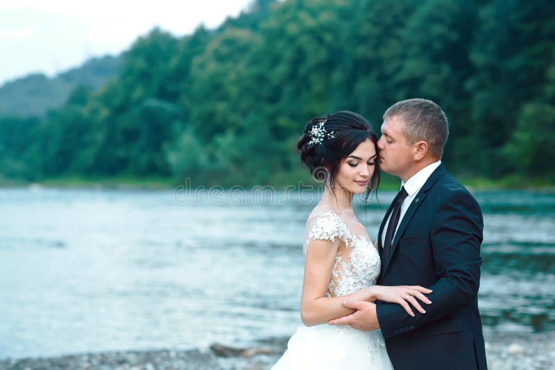 Couples romantiques de nouveaux mariés étreignant près du lac bleu, jeune mariée magnifique de embrassement de marié sensuel par photos stock Couples romantiques de nouveaux mariés étreignant près du lac bleu, jeune mariée magnifique de embrassement de marié sensuel par photos stock