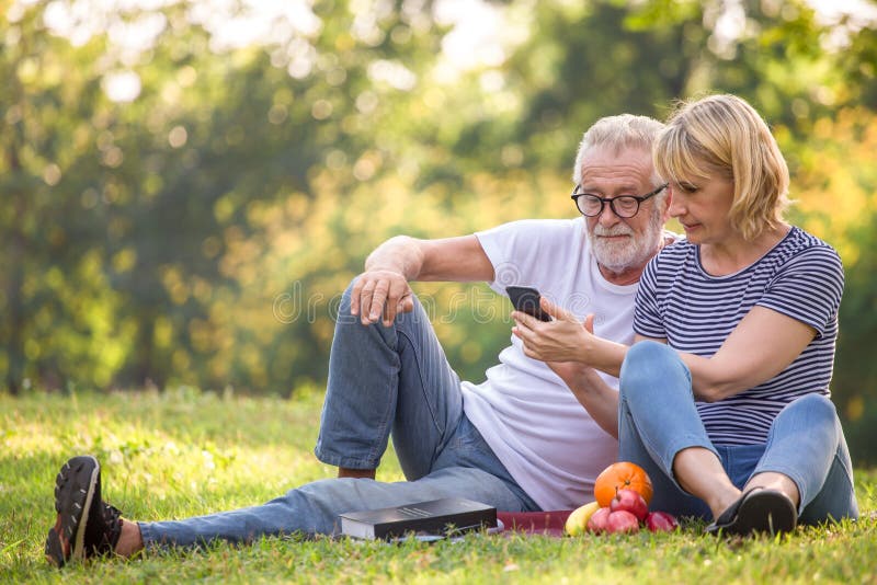 Couples supérieurs heureux détendant en parc utilisant le smartphone ensemble personnes âgées s'asseyant sur l'herbe en parc d'ét image stock