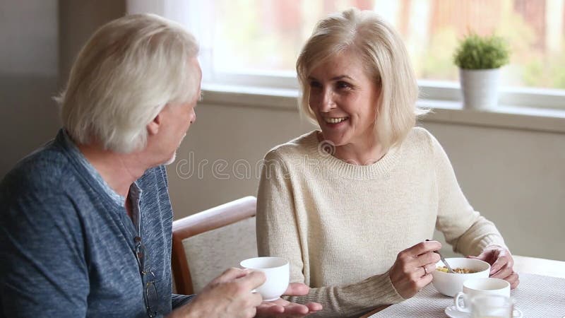 Couples âgés moyens heureux parlant mangeant du café potable de petit déjeuner ensemble banque de vidéos