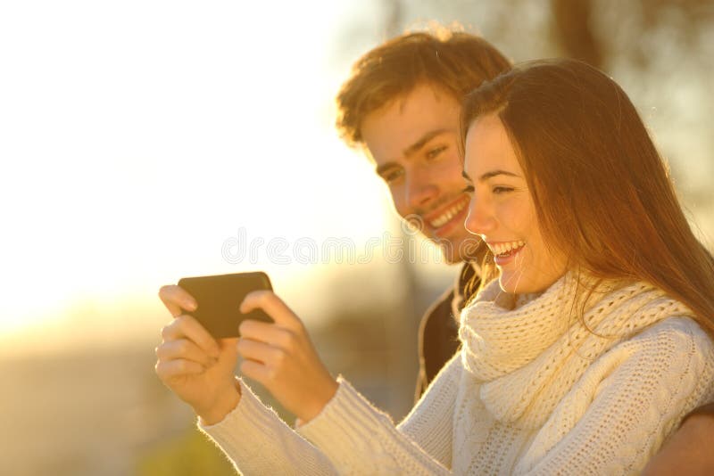 Happy couple watching media videos in a smart phone at sunset on the beach. Happy couple watching media videos in a smart phone at sunset on the beach