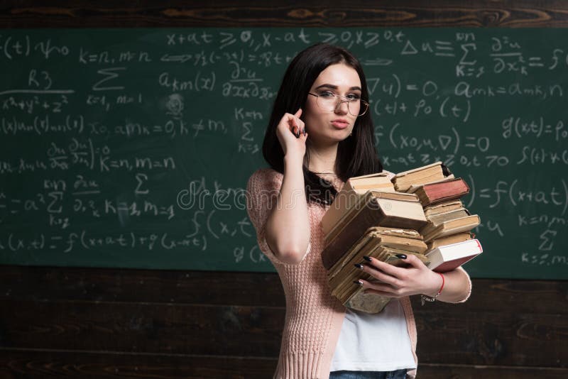 Course project. Girl holds heavy pile of old books, chalkboard background. Girl student works on scientific research.