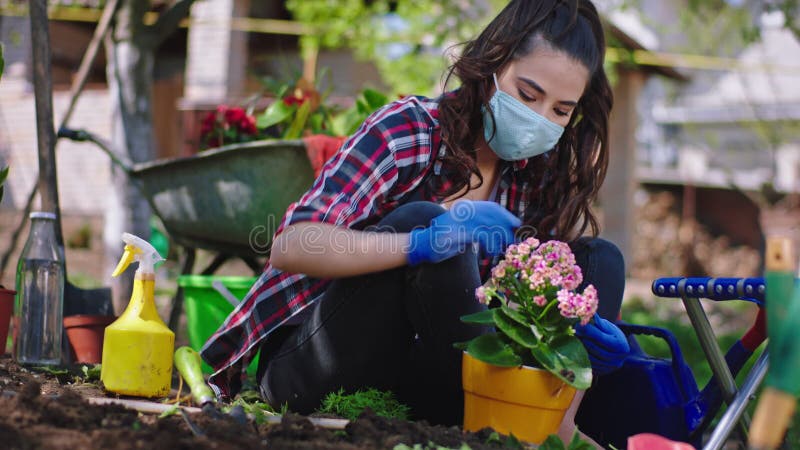 Covid19 concept lady avec masque de protection dans le jardin prendre soin des fleurs de la marmite qu'elle a concentrée banque de vidéos