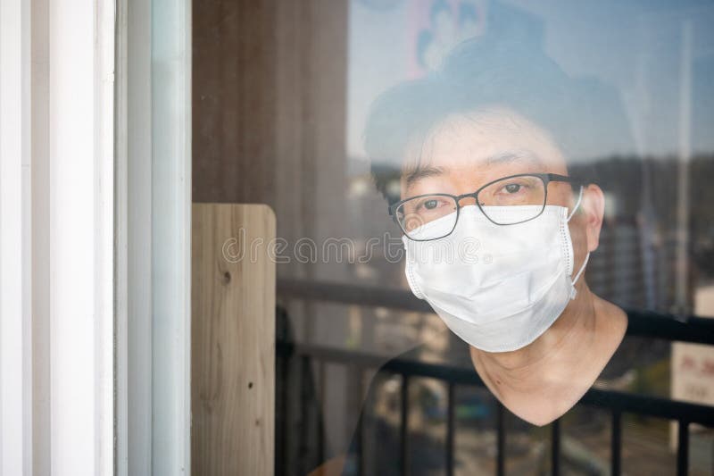 COVID-19 coronavirus self-isolation concept. An Asian man with a face mask and a window looking out the window during his self-