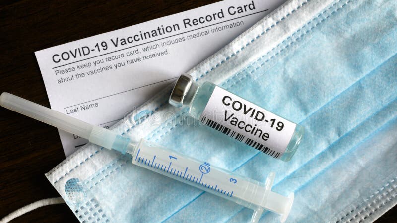 COVID-19 vaccine bottle, syringe, surgical mask and coronavirus Vaccination Record Card on desk, top view of corona virus vaccine and immunization certificate required for travel due to COVID pandemic. COVID-19 vaccine bottle, syringe, surgical mask and coronavirus Vaccination Record Card on desk, top view of corona virus vaccine and immunization certificate required for travel due to COVID pandemic