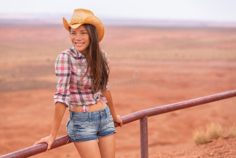 Cowgirl woman smiling happy on country farm landscape wearing cowboy hat and western shirt and jeans shorts. Young