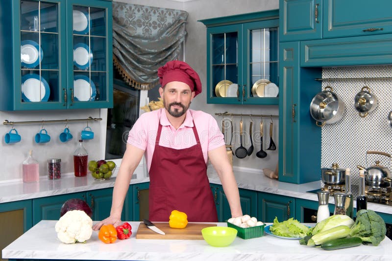 Cozinheiro chefe farpado envelhecido no avental vermelho e no tampão vermelho que estão na cozinha t foto de stock Cozinheiro chefe farpado envelhecido no avental vermelho e no tampão vermelho que estão na cozinha t foto de stock