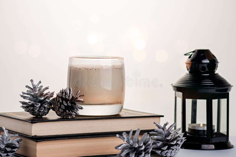 Cozy ainda vive com a bebida tradicional de ovos em pilhas de livros com decoração de pínus prateados e vela a queimar imagens de stock Cozy ainda vive com a bebida tradicional de ovos em pilhas de livros com decoração de pínus prateados e vela a queimar imagens de stock