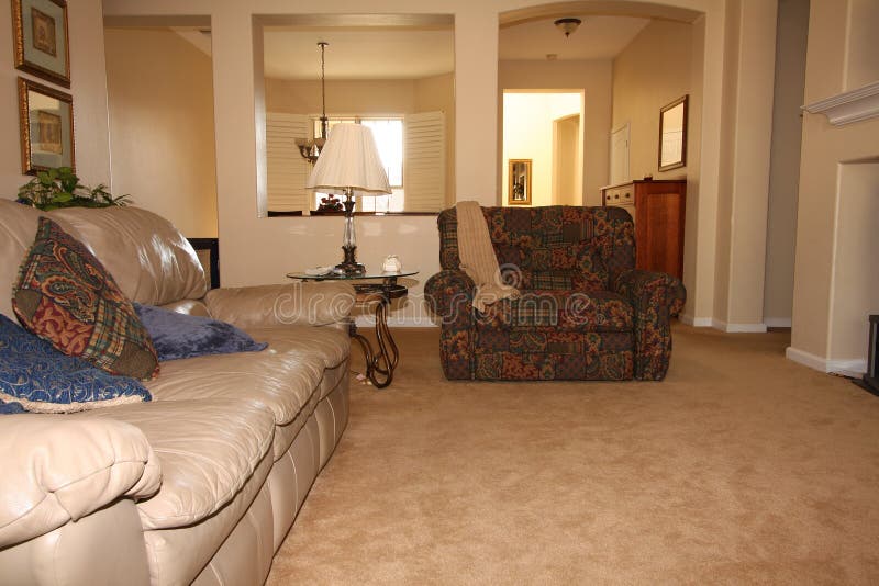 A neutral colored living room in a big modern house has beige carpet, a beige leather couch, patterned loveseat, glass end table with lamp, and interesting architecture. A neutral colored living room in a big modern house has beige carpet, a beige leather couch, patterned loveseat, glass end table with lamp, and interesting architecture.