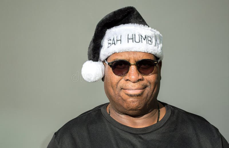 Cranky middle aged rotund African American man wearing a black and white Santa hat saying Bah Humbug against a solid background. Cranky middle aged rotund African American man wearing a black and white Santa hat saying Bah Humbug against a solid background