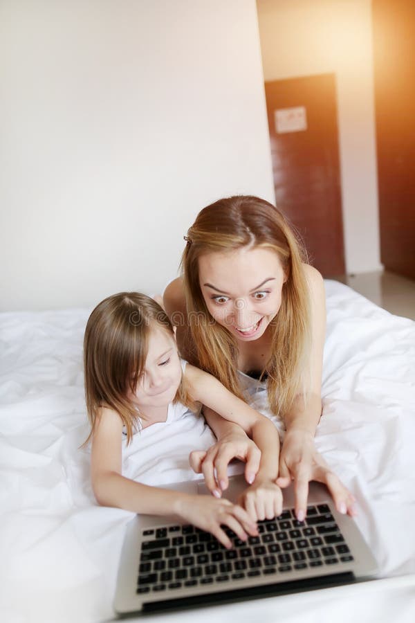 Crazy happy mother play games with little daughter at laptop laying on white bed with sunshine royalty free stock photography Crazy happy mother play games with little daughter at laptop laying on white bed with sunshine royalty free stock photography