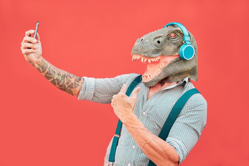 Crazy senior man wearing t-rex mask and taking selfie with mobile smartphone - Hipster older male having fun listening music