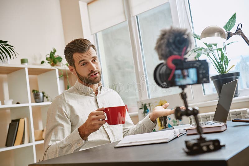 Creating content. Cheerful male blogger making a new video for his vlog and drinking a tea while sitting indoors. royalty free stock photography Creating content. Cheerful male blogger making a new video for his vlog and drinking a tea while sitting indoors. royalty free stock photography