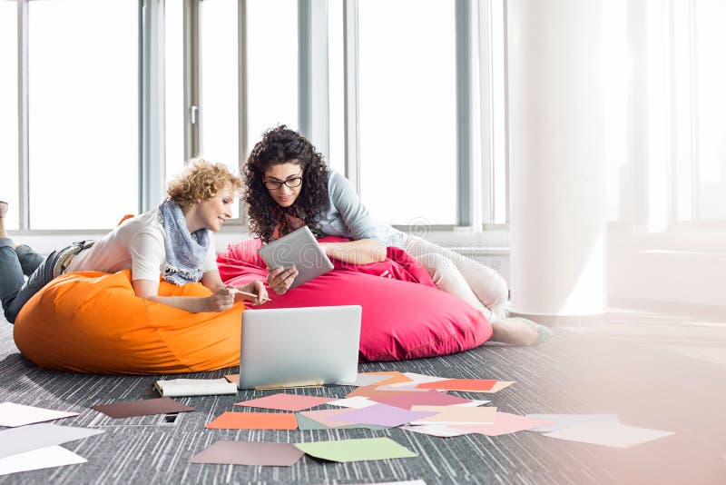 Creative businesswomen using tablet PC while relaxing on beanbag chairs at office