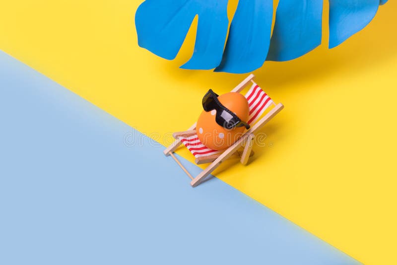 Creative funny composition with Easter egg with sunglasses sitting on deck chair and blue monstera leaf on illuminating yellow and royalty free stock photo