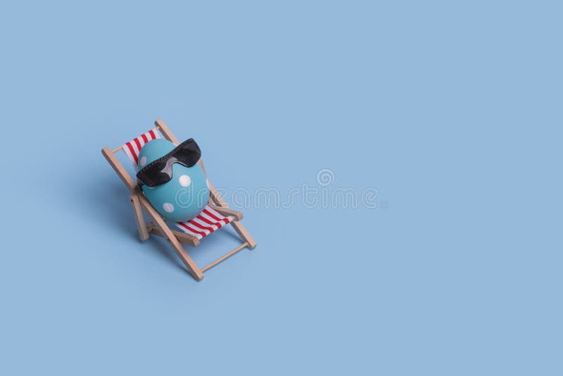 Creative funny composition with Easter egg with sunglasses while sitting on deck chair on bright blue background royalty free stock photo
